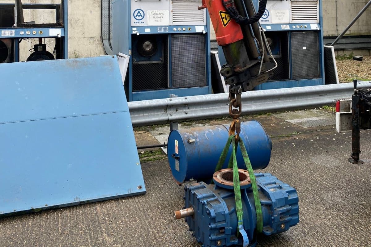 Blower stage on lifting straps below a crane hook beside two aeration blowers